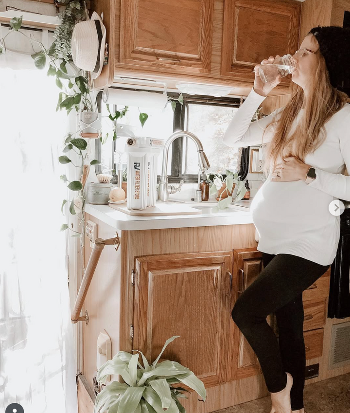 Woman drinking water in a cozy kitchen with wooden cabinets and plants.